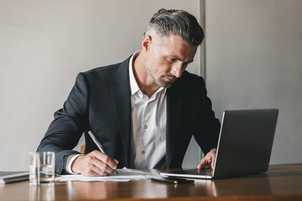 Bild eines konzentrierten selbstbewussten Geschäftsmannes in weißem Hemd und schwarzem Anzug, der im Büro am Laptop arbeitet und Notizen aufschreibt - Foto, Bild