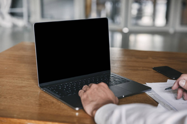 Cropped image of businesslike man wearing white shirt and black suit sitting at table in office and looking at copyspace screen of laptop during work with documents - Foto, Bild