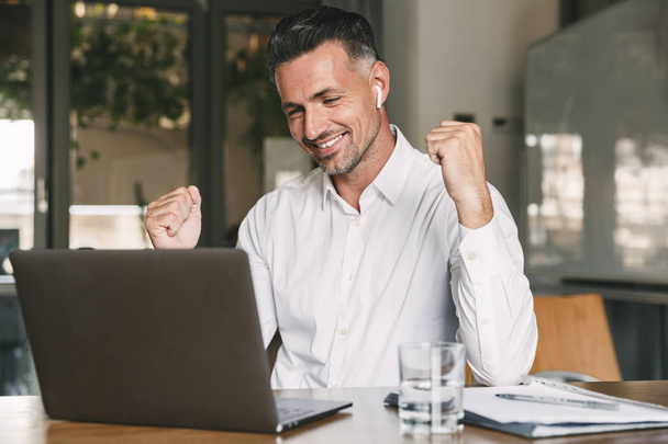 Photo of happy businessman 30s wearing white shirt and wireless earphones rejoicing and clenching fist while working on laptop in office - Foto, Imagen