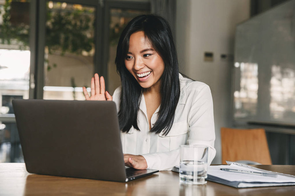 Image of happy asian woman 20s wearing white shirt smiling and waving hand at laptop while speaking or chatting on video call in office - Valokuva, kuva