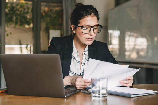 Photo of serious manager or director woman wearing formal clothing and eyeglasses holding in hands paper documents while working in office on laptop - Photo, Image