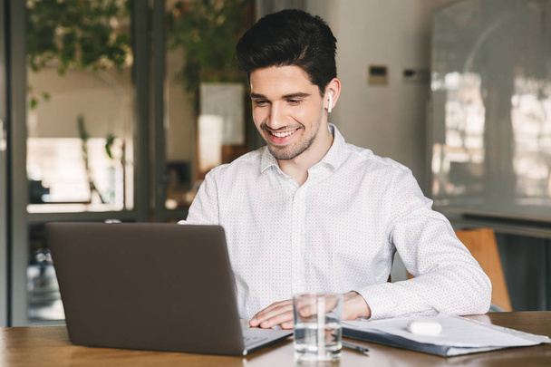 Изображение красивого бизнесмена в белой рубашке и современной наушнике, улыбающегося сидя за столом в офисе и глядя на ноутбук
 - Фото, изображение