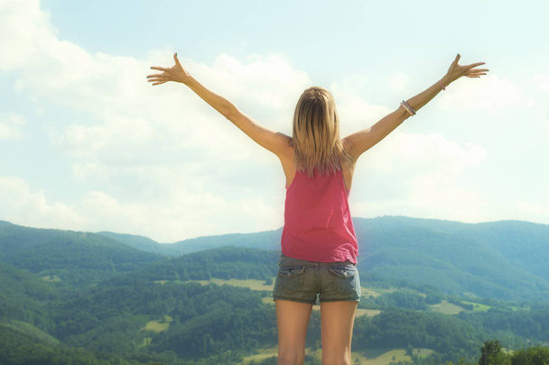 вид сзади женщины с поднятыми руками на зеленом холме в солнечный день
  - Фото, изображение