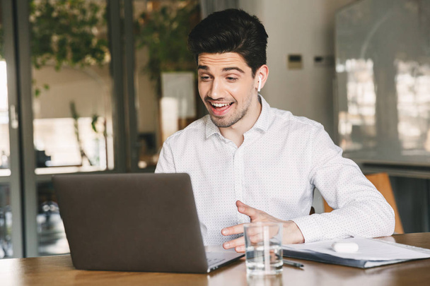 Image of cheerful office man wearing white shirt and modern earpod smiling while sitting at table in office and speaking to laptop during video call or chat - Fotoğraf, Görsel
