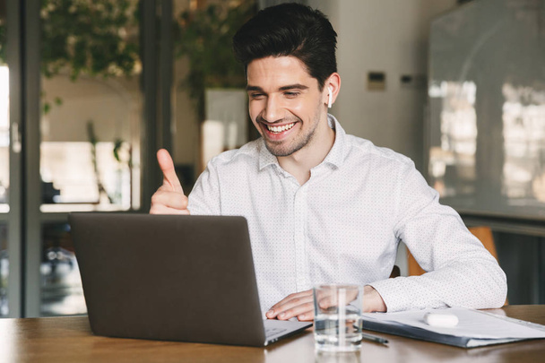 Portrait of successful businessman 30s wearing white shirt laughing and showing thumb up at laptop in office during video conference or call using bluetooth earbud - Фото, изображение