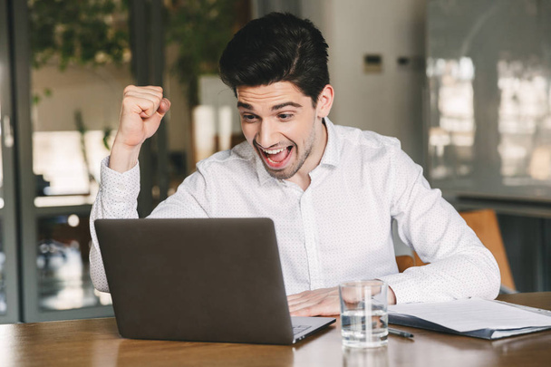 Photo of excited young man 30s wearing white shirt and bluetooth earbuds screaming and clenching fist like winner while looking at laptop in office - Fotografie, Obrázek