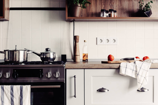 Cozinhar na cozinha moderna em estilo escandinavo. elegante cozinha interior com mobiliário moderno e aparelhos de aço inoxidável. bancada de madeira, fogão de aço, placa, faca e especiarias, tomates
 - Foto, Imagem