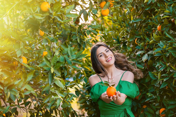 fröhliche junge Frau, draußen bei Sonnenuntergang in einem orangefarbenen Obstgarten, in die Kamera blickend und lächelnd, eine orangefarbene Frucht in der Hand. Konzept für gesunden Lebensstil, Haut- und Haarpflege. - Foto, Bild