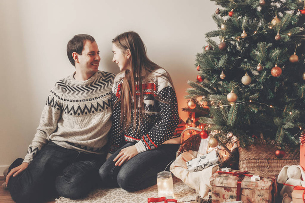 Stilvolles Paar in Pullovern umarmt und lächelnd am Weihnachtsbaum im gemütlichen Abendzimmer. Stimmungsvolle Momente. Frohe Weihnachten und ein gutes neues Jahr. frohe Feiertage - Foto, Bild