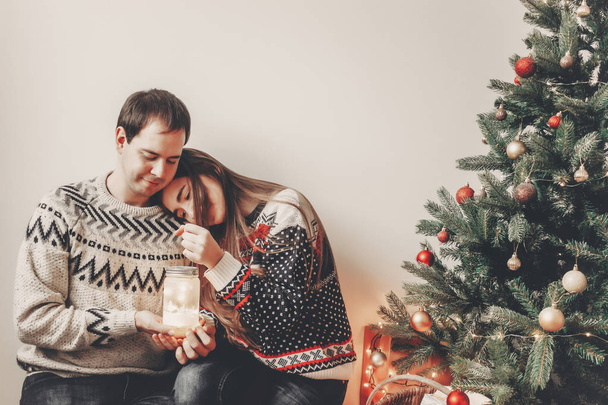 happy couple in stylish sweaters holding lantern light in festive room at christmas tree and embracing. atmospheric festive moments. merry christmas and happy new year concept - Photo, Image