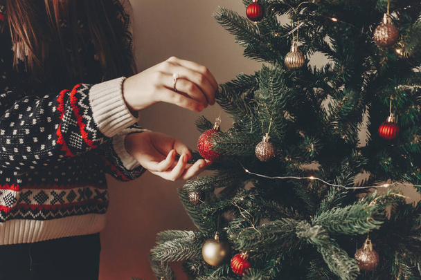 woman in stylish sweater decorating christmas tree in room. merry christmas and happy new year concept. space for text. woman putting on red ornaments. family cozy moments - Fotó, kép