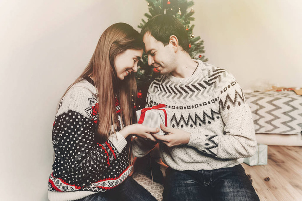 メリー クリスマスと新年あけましておめでとうございます概念。ライトとクリスマス ツリーでお祭り部屋にギフトと赤の弓を保持のセーターでスタイリッシュな流行に敏感なカップル。楽しい休暇をお過ごしください。家族の瞬間 - 写真・画像