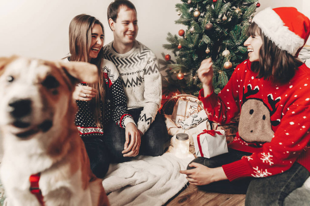 ευτυχισμένη οικογένεια σε κομψό πουλόβερ και χαριτωμένο αστείο σκύλο ανταλλάσσοντας δώρα στο χριστουγεννιάτικο δέντρο με φώτα. συναισθηματικές στιγμές. καλά Χριστούγεννα και Ευτυχισμένο το νέο έτος έννοια. χώρο για το κείμενο - Φωτογραφία, εικόνα