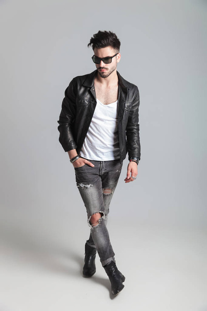 man with leather jacket and grey jeans looks to side while standing on light grey background with hand in pocket, full length picture - Φωτογραφία, εικόνα