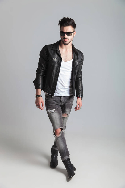 handsome man wearing leather jacket and sunglasses stands on light grey background cross-legged and looks to side - Фото, изображение