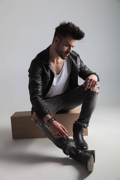 young man in leather jacket and grey ripped jeans sitting on a wooden box and looking down to side, on light grey background - Фото, изображение