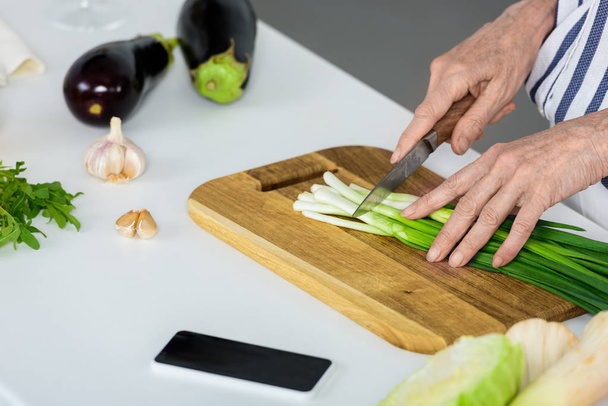 imagen recortada de mujer de pelo gris cortando cebolla verde en tablero de madera en la cocina
  - Foto, Imagen
