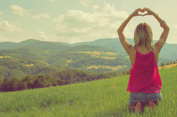 zadní pohled na ženu s rukou ukazující srdce znamení sedíte na kolenou v zelený trávník za slunečného dne  - Fotografie, Obrázek