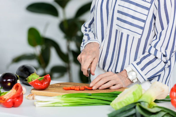 cropped image of senior woman cutting red bell pepper on wooden board in kitchen  - Foto, imagen
