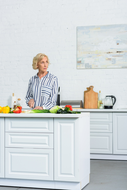 beautiful grey hair woman cutting vegetables in kitchen and looking away  - Foto, afbeelding