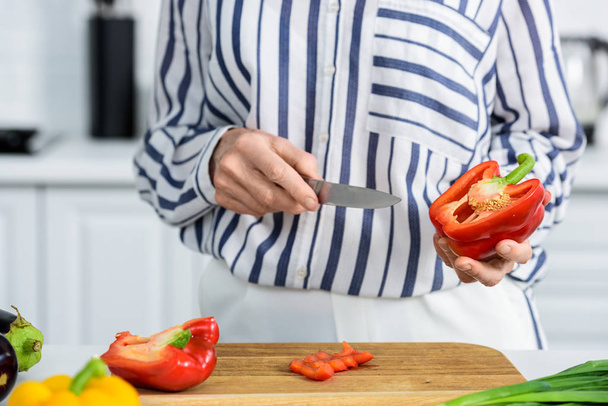 abgeschnittenes Bild einer grauhaarigen Frau, die in der Küche rote Paprika auf einem Holzbrett schneidet  - Foto, Bild