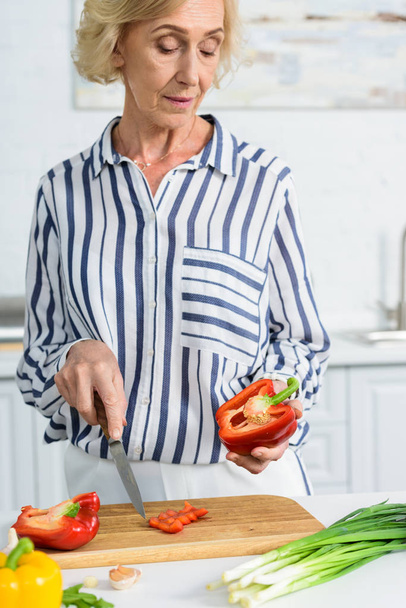 attractive grey hair woman cutting red bell pepper on wooden board in kitchen  - Fotografie, Obrázek