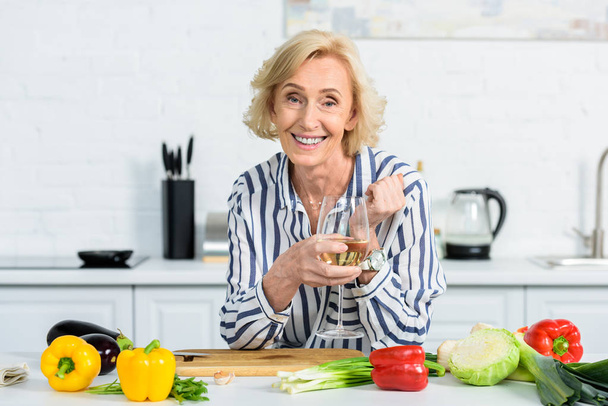 şarap tutarak ve mutfak kamerada bakarak mutlu çekici gri saçlı kadın  - Fotoğraf, Görsel