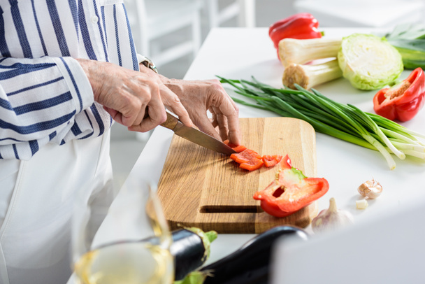 image recadrée d'une femme âgée coupant du poivron rouge sur une planche en bois dans la cuisine
  - Photo, image