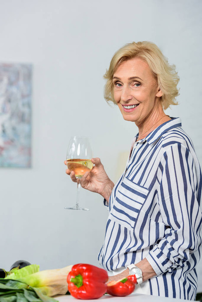 smiling attractive senior woman holding glass of white wine in kitchen and looking at camera - Fotó, kép