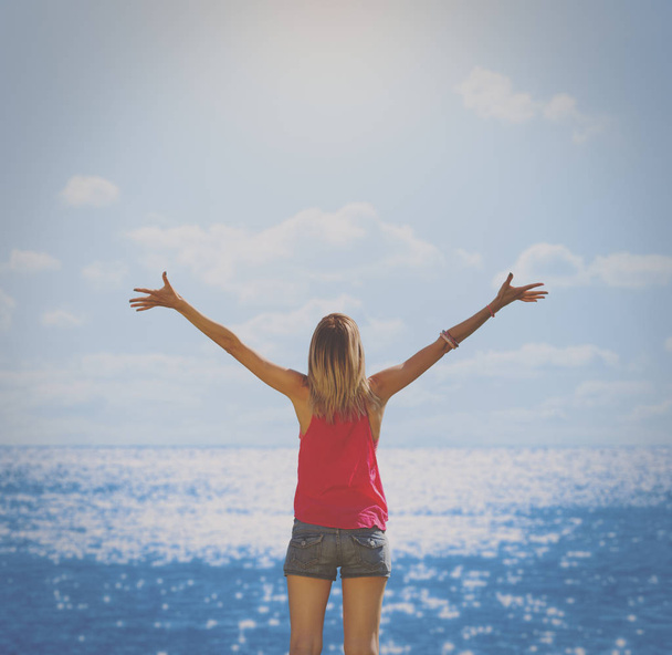 takaisin näkymä nainen seisoo nosti kätensä meren rannalla aurinkoisena aamuna
  - Valokuva, kuva