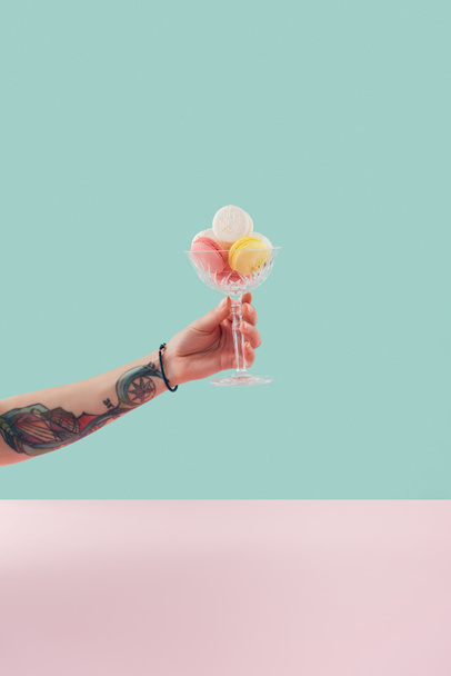 cropped view of tattooed hand with macarons in glass bowl on pastel background - Zdjęcie, obraz
