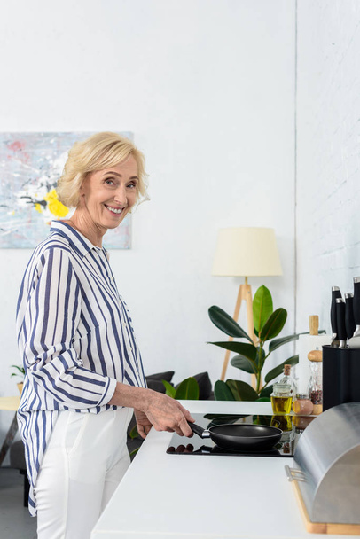 souriant attrayant cheveux gris femme cuisine et mettre la poêle sur la cuisinière
  - Photo, image