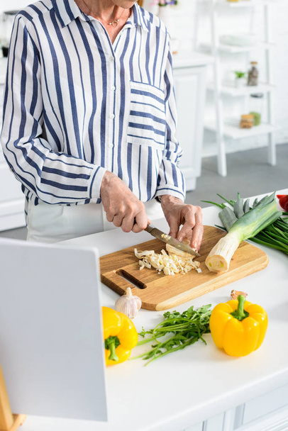 cropped image of senior woman cutting uncooked leek on wooden board in kitchen  - Fotó, kép
