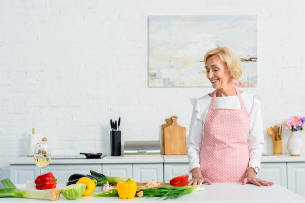 lachende aantrekkelijke senior vrouw permanent op de aanrecht en groenten te kijken - Foto, afbeelding