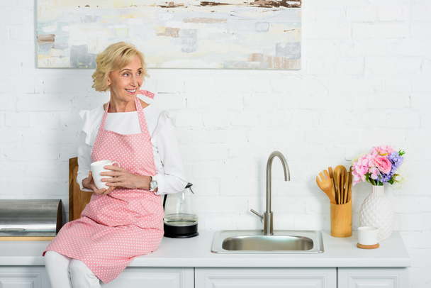 приваблива старша жінка сидить на кухонній стійці з чашкою кави і дивиться в сторону
 - Фото, зображення