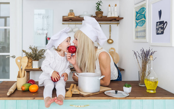 Mère caucasienne et petite fille en bonnet blanc mangent des pommes dans la cuisine. Concept de nutrition saine
. - Photo, image