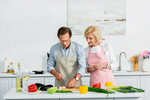 ανώτερων γυναίκα ψάχνει άντρα πώς κοπής λαχανικών στην κουζίνα - Φωτογραφία, εικόνα