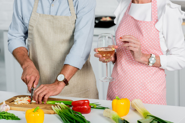 geschnittenes Bild von älterem Mann, der Gemüse schneidet und Frau, die ein Glas Wein in der Küche hält - Foto, Bild
