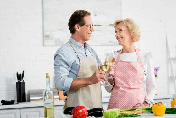 happy senior couple clinking with glasses of wine during cooking at kitchen and looking at each other - Фото, зображення