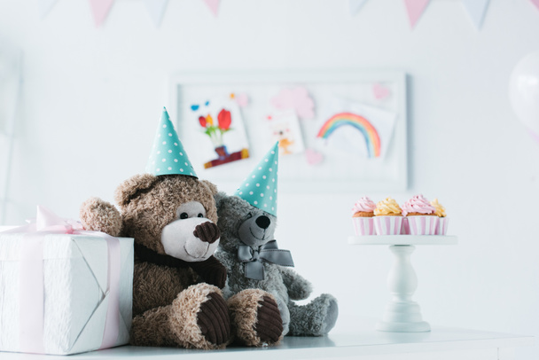 teddy bears in cones on table with cupcakes on stand and present box  - Фото, изображение