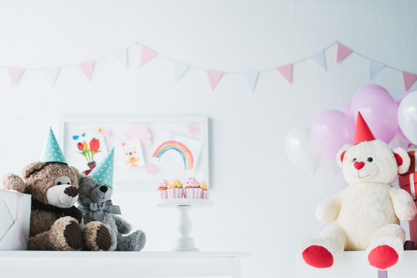 teddy bears in cones on table with cupcakes on stand in decorated for birthday room - Fotó, kép