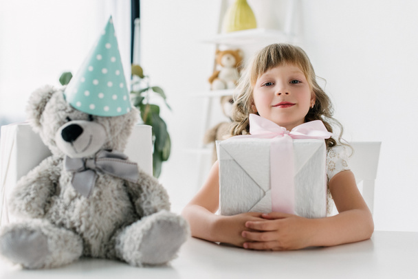 happy birthday kid sitting with present at table with teddy bear in cone - Photo, Image
