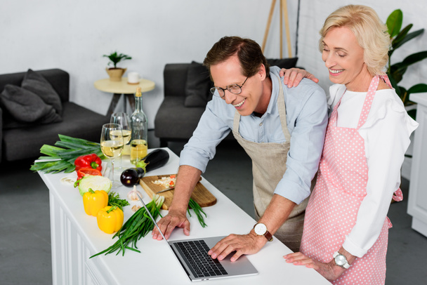 hymyilevä vanhempi pari ruoanlaitto keittiössä ja kannettavan tietokoneen avulla
 - Valokuva, kuva