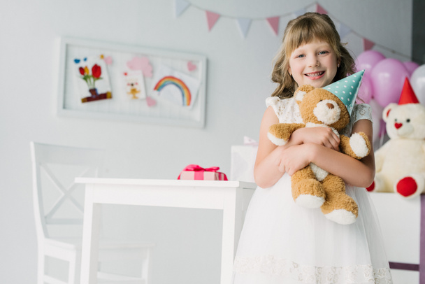adorable birthday kid looking at camera and holding teddy bear in cone  - Photo, Image