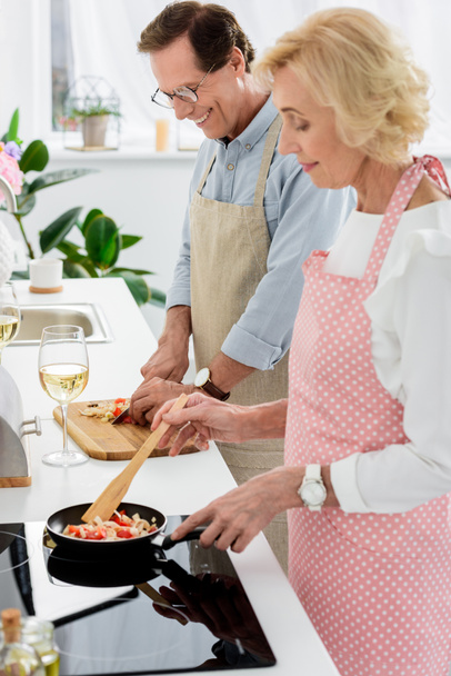 side view of senior couple cooking vegetables on frying pan and cutting vegetables on wooden board at kitchen - Foto, Bild
