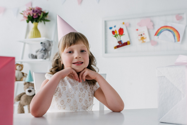 smiling birthday child in cone looking at camera and sitting at table with gifts  - Foto, imagen