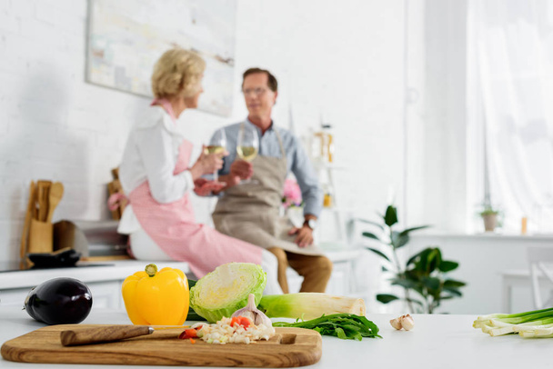close-up view of fresh vegetables in wooden cutting board and senior couple drinking wine behind in kitchen - Photo, Image
