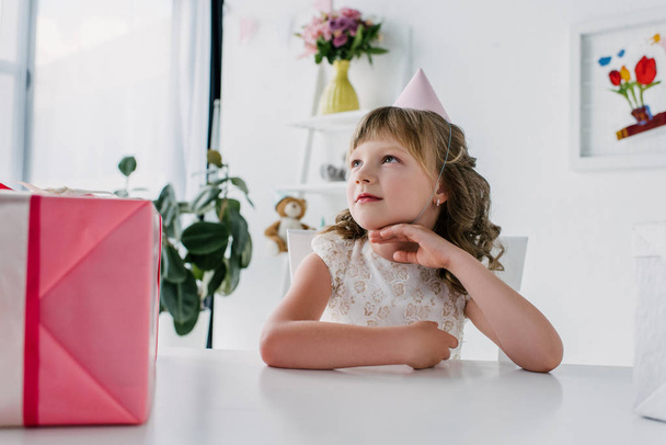 thoughtful birthday kid looking away and sitting at table with present  - Foto, imagen