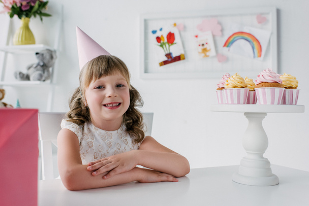 lindo cumpleaños niño en cono mirando a la cámara y sentado en la mesa con cupcakes
  - Foto, Imagen