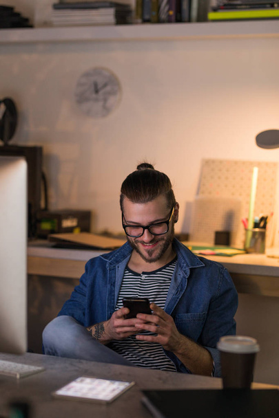 Kaukaski młody, uśmiechnięty człowiek siedzący projektanta w jego biurze w godzinach wieczornych i pisania na telefon komórkowy. - Zdjęcie, obraz